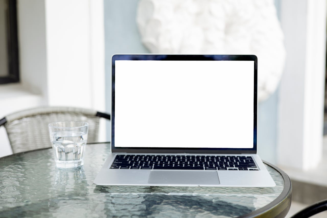 Image of an open laptop with a blank screen on a glass table, next to a glass of water.